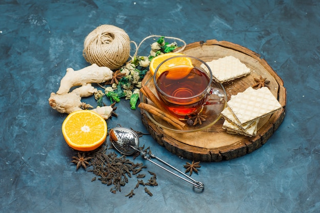 Tea with herbs, orange, spices, waffle, thread, strainer in a mug on wooden board and stucco background, flat lay.