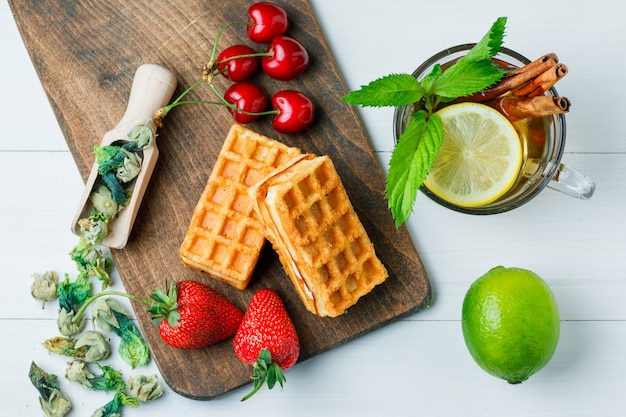 Tea with lemon, waffle, dried herbs, fruits, mint, cinnamon in a cup on wooden and cutting board, flat lay.