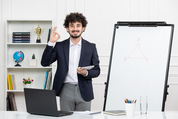 Teacher brunette instructor with computer in suit and whiteboard in classroom showing ok sign