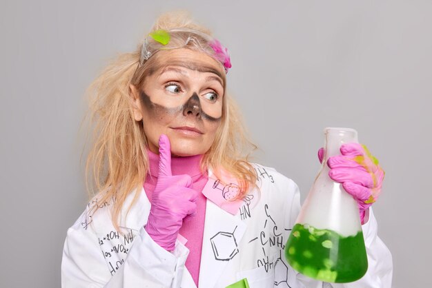 Teacher of chemist looks attentively at glass beaker with green liquid works in chemistry lab wears rubber gloves and medical coat isolated over grey background. Science and research concept