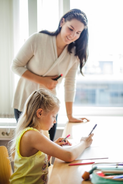 Teacher leaning upon table watching schoolgirl writing