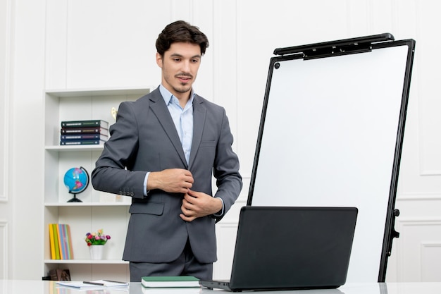 Teacher smart instructor in grey suit in classroom with computer and whiteboard buttoning suit