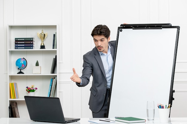 Teacher smart instructor in grey suit in classroom with computer and whiteboard explaining a task