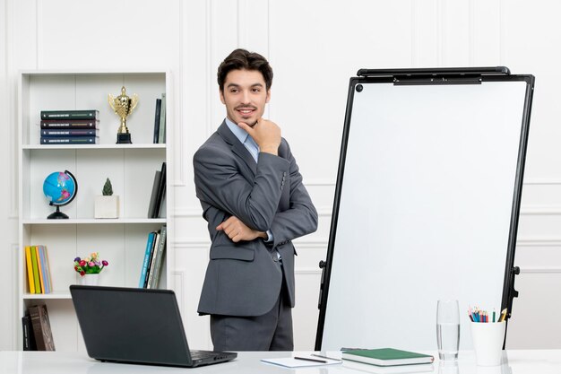 Teacher smart instructor in grey suit in classroom with computer and whiteboard looking confident