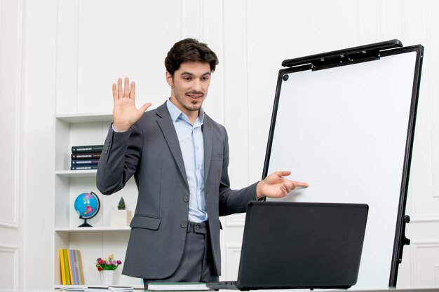 Teacher smart instructor in grey suit in classroom with computer and whiteboard saying goodbye