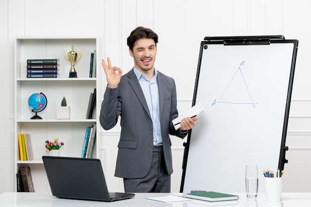 Teacher smart instructor in grey suit in classroom with computer and whiteboard showing ok sign