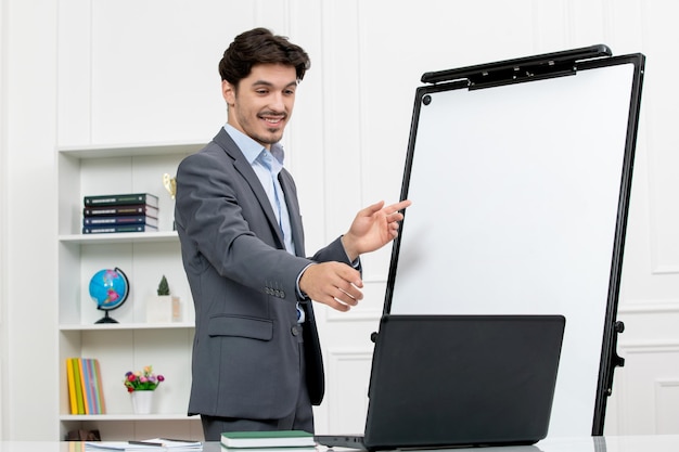 Teacher smart instructor in grey suit in classroom with computer and whiteboard smiling on video