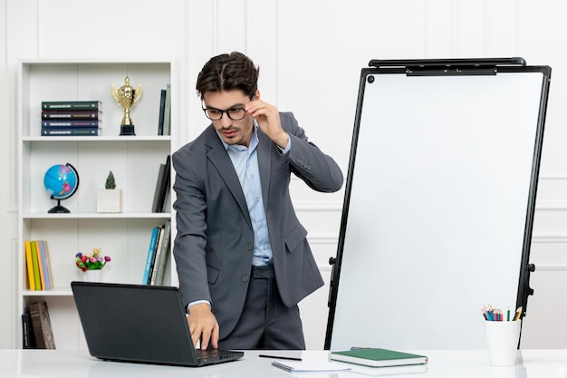 Teacher smart instructor in grey suit in classroom with computer and whiteboard touching glasses