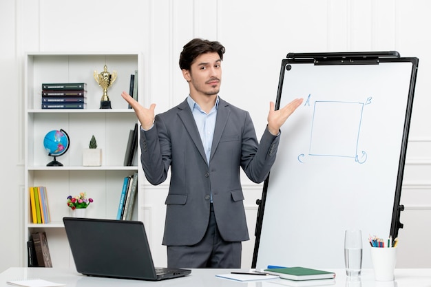 Teacher smart instructor in grey suit in classroom with computer and whiteboard waving hands