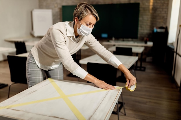 Free photo teacher with face mask preparing classroom and marking tables with a tape