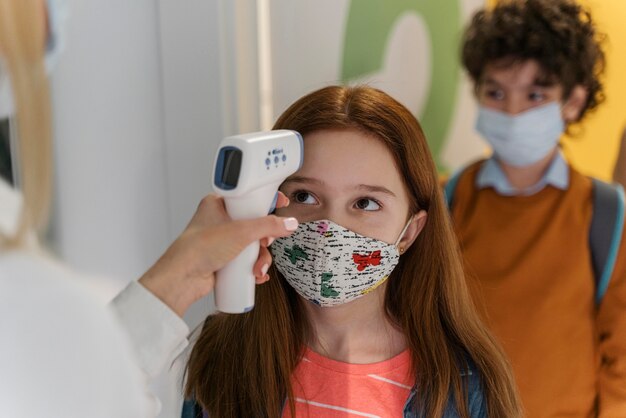 Teacher with medical mask checking children's temperature in school