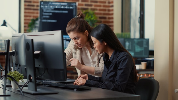 Team of coworkers comparing source codes running on laptop screen and computer monitor in it development office. software developers collaborating on data coding group project while sitting at desk.