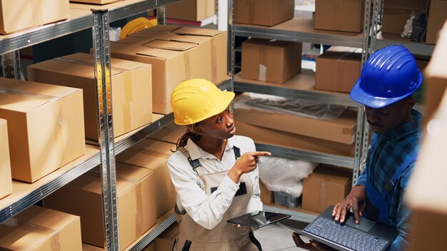 Team of employees checking stock on laptop and tablet, using devices to plan goods logistics in warehouse area. Young people working with cardboard packages and cargo products. Tripod shot.