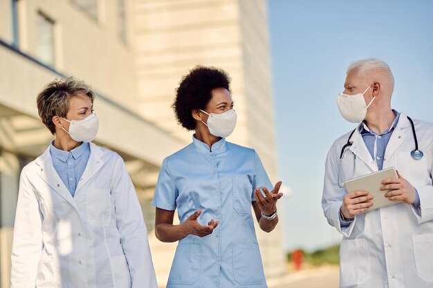 Team of happy doctors communicating while walking outdoors with protective masks on their faces