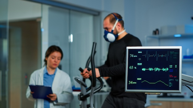 마스크 런닝을 착용한 남자 퍼포먼스 스포츠의 vo2를 모니터링하는 중간 연구원 팀. Ekg 스캔이 실험실의 컴퓨터 화면에서 실행되는 동안 스포츠맨의 지구력을 측정하는 실험실 과학 의사