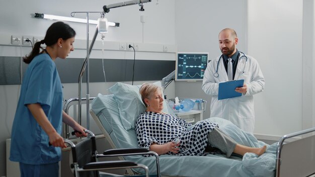 Team of nurse and doctor giving assistance to ill woman with wheelchair and transportation from hospital ward bed. Medical workers helping retired patient after healthcare consultation