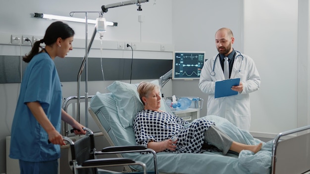 Team of nurse and doctor giving assistance to ill woman with wheelchair and transportation from hospital ward bed. Medical workers helping retired patient after healthcare consultation