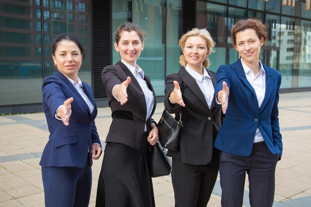 Team of positive successful businesswomen standing together near office building, offering handshake, looking at camera. Front view. Cooperation concept