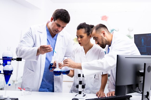 흰색 코트를 입은 과학자 팀이 현대 연구실에서 파란색 액체를 피우며 함께 일하고 있습니다.