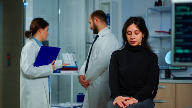 Команда ученых-врачей обсуждает состояние здоровья пациента, функции мозга, нервную систему, томографическое сканирование, пока женщина ждет диагноза заболевания, сидя в неврологической исследовательской лаборатории
