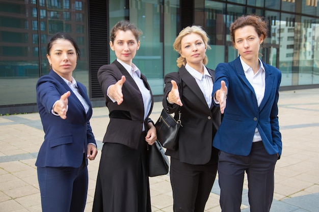 Team of serious confident businesswomen standing together near office building, offering handshake, looking at camera. Front view. Cooperation concept