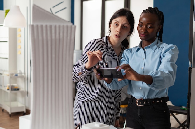 Team of two architects working with smartphone to find inspiration in front of foam scale model maquette of skyscraper. Engineers with mobile device next to desk in architectural modern office.