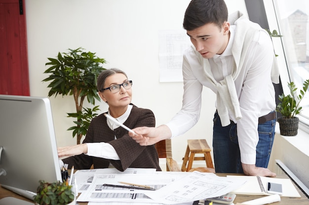 Team of two skillful architects young man and elderly woman developing new residential housing project, working on generic computer in office, using CAD program, man pointing at screen with pencil