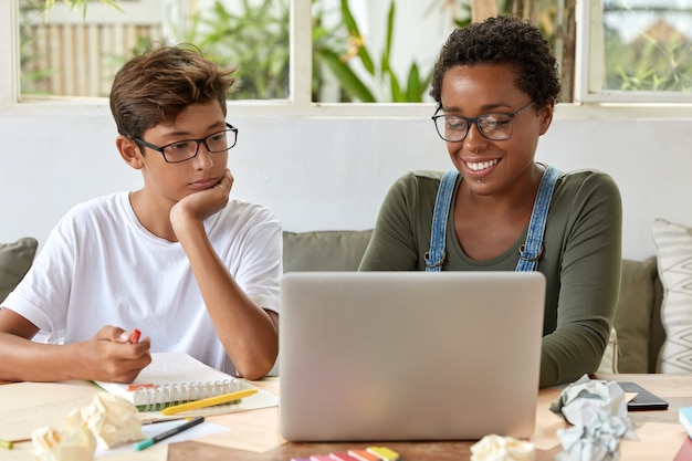 Team work concept. Black clever female teacher and her pupil watch training video together, focused into laptop computer, connected to internet, sit at desktop with notepad for making records