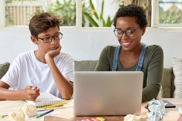 Концепция работы в команде. Черная умная учительница и ее ученица вместе смотрят обучающее видео, сосредоточены на портативном компьютере, подключены к Интернету, сидят за рабочим столом с блокнотом для записи