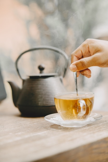 テーブルの上のティーポットと熱いお茶のカップ。小さじ1杯を持っている手