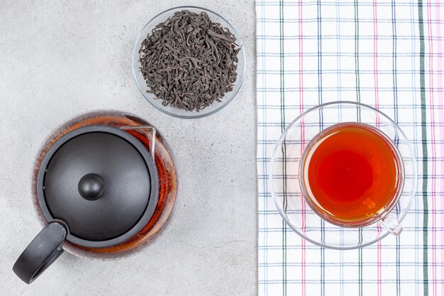 A teapot and a small bowl of leaves next to a cup of tea on towel on marble background. High quality photo