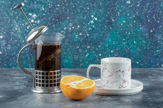 Чайник с чашкой чая и свежим лимоном над серым столом.