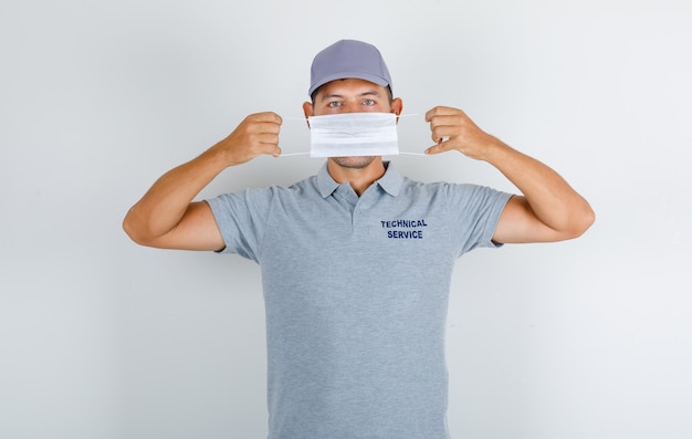 Человек технической службы в медицинской маске в серой футболке с кепкой и осторожно смотрит
