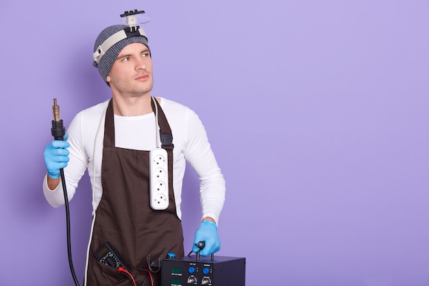 Free photo technician being ready to solder something, attractive male wears white casual shirt, cap and brown apron, holds soldering iron