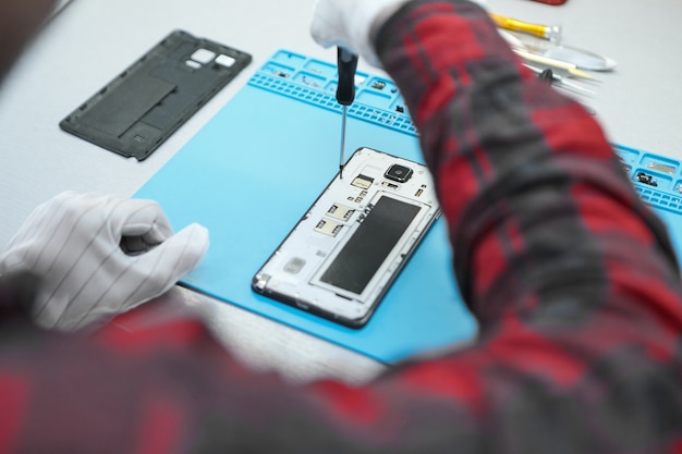 Technician wearing white antistatic gloves and plaid shirt sitting at his desk and using precision screwdriver to remove screws on back of faulty mobile phone