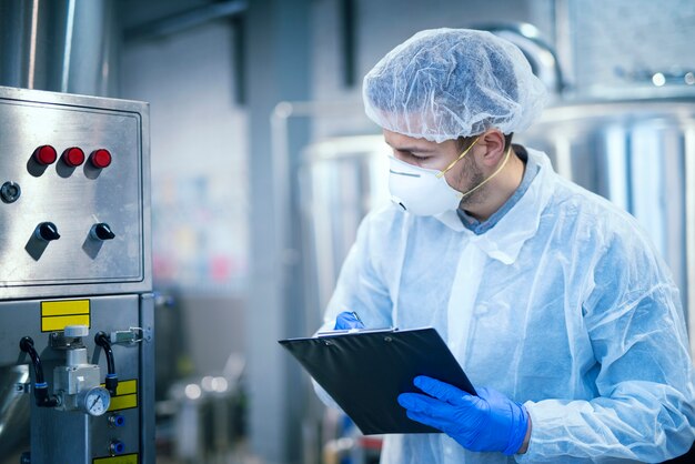 Technologist expert in protective uniform with hairnet and mask taking parameters from industrial machine in food production plant