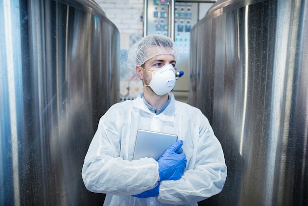 Technologist in protective uniform with tablet standing by metal reservoir in food processing industry