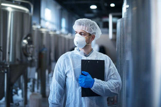 Technologist in protective white suit with hairnet and mask standing in food factory