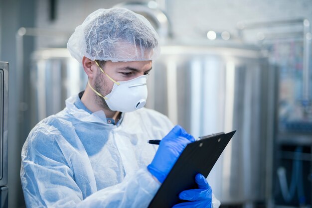 Technologist specialist with protective mask and hairnet holding clipboard checklist and writing production results