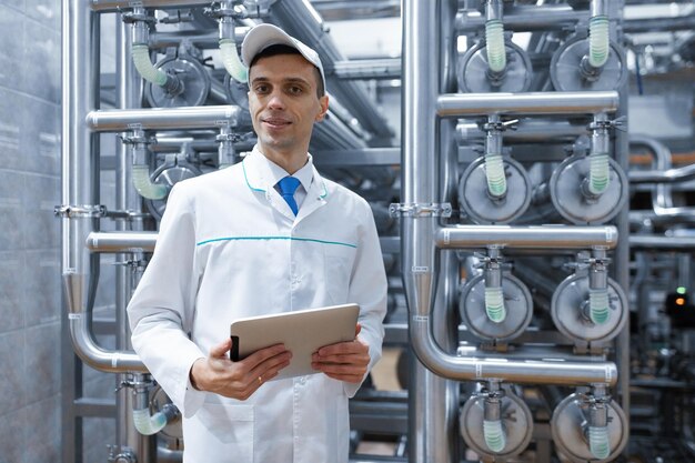 Technologist in a white coat makes the necessary entries in the tablet is at the factory A man with a digital tablet in uniform in the production shop interior of production department on background