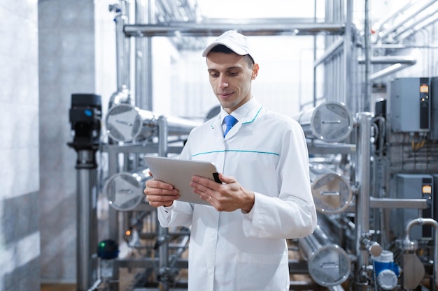 Technologist in a white coat makes the necessary entries in the tablet is at the factory A man with a digital tablet in uniform in the production shop interior of production department on background