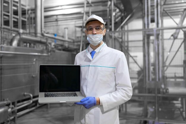 노트북을 손에 들고 흰 코트를 입은 기술자는 낙농장에서 생산 공정을 제어합니다. 글을 쓰는 장소는 노트북 컴퓨터가 있는 기술자가 공장에 있습니다