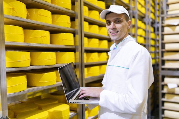 노트북을 손에 들고 흰 코트를 입은 기술자가 버터와 치즈 생산을 위한 가게의 치즈 창고에 있습니다. 낙농 공장의 품질 관리 치즈가 있는 랙