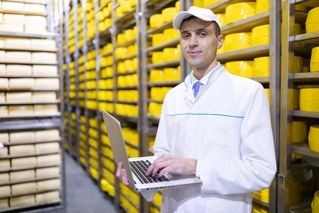 Technologist in a white coat with a laptop in his hands is in the warehouse of cheese in the shop for the production of butter and cheese Quality control at the dairy plant Racks with cheese