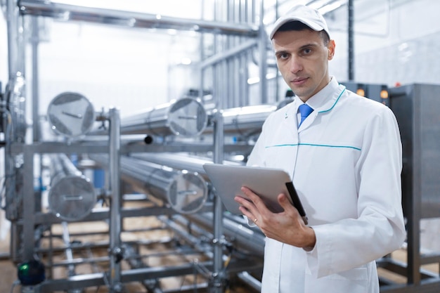 Technologist in a white coat with a tablet in his hands controls the production process in the dairy shop Quality control at the dairy plant