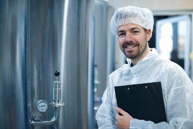 Free photo technologist in white protective uniform and hairnet standing by chrome reservoirs with pressure gauge in food processing plant