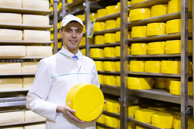 Technologist in a white robe with a yellow cheese head in his hands is in the shop for the production of butter and cheese The production process at the plant of dairy products Racks with cheese