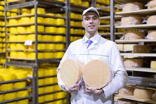 Technologist in a white robe with a yellow cheese head in his hands is in the shop for the production of butter and cheese The production process at the plant of dairy products Racks with cheese