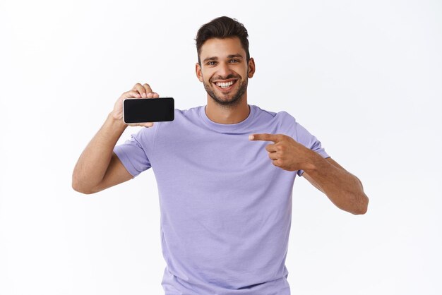 기술 광고 및 가제트 개념 스마트폰을 수평으로 가리키는 모바일 디스플레이를 들고 멋진 게임을 보여주는 보라색 티셔츠를 입은 행복한 미소 힙스터 남자