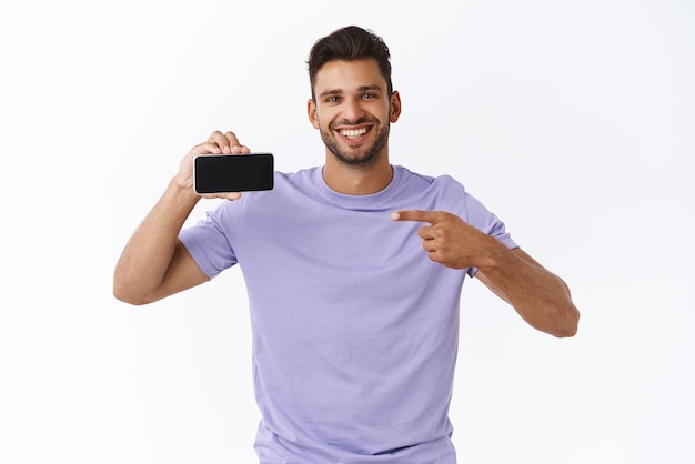 Технологическая реклама и концепция гаджетов Счастливый улыбающийся хипстер в фиолетовой футболке с щетиной, показывающий потрясающую игру со своим собственным счетом, держа смартфон горизонтально, указывая на мобильный дисплей